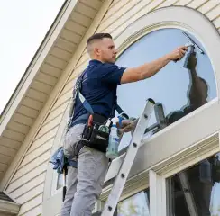 Professional window cleaner on a ladder using a squeegee to clean an exterior arched window, Fort Worth, TX.